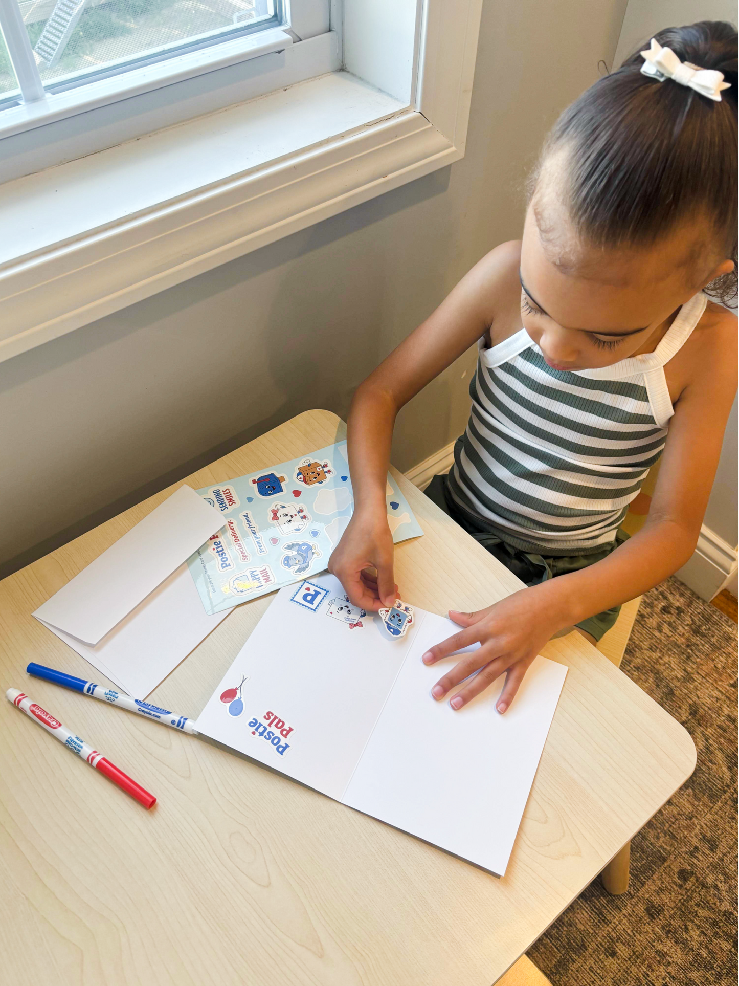 A young girl with her hair in a bow sits at a light wood table, placing stickers on a Postie Pals Starter Card.